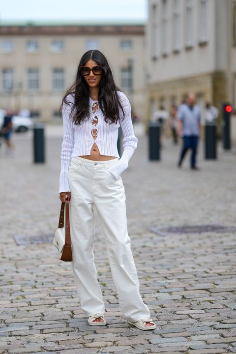 a woman wearing a white tie front cardigan with low rise white jeans, flat comfortable sandals, and a two tone brown and white bag on the street during copenhagen fashion week in a roundup of cute summer outfits 2023