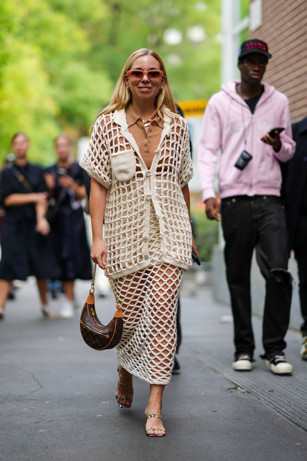 a woman wearing slim tan rectangular sunglasses, necklaces, earrings, a camel knit top, an open weave cream color large fishnet crochet jacket, a matching midi skirt, a louis vuitton bag, and snakeskin strappy sandals in a roundup of the best travel outfits for women 2024