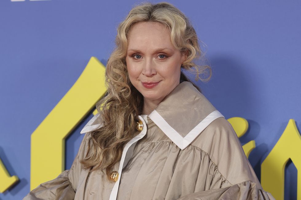 gwendoline christie, a woman looks at the camera and smiles slightly, curled blonde hair falls over right shoulder, wearing a brown oversized dress