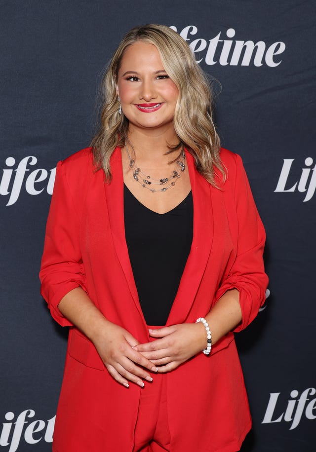 gypsy rose blanchard smiles at the camera, she wears a red suit jacket and black top and stands in front of a black background