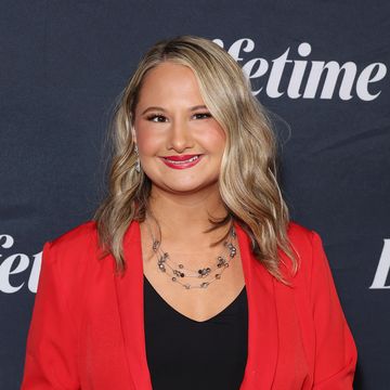 gypsy rose blanchard smiles at the camera, she wears a red suit jacket and black top and stands in front of a black background