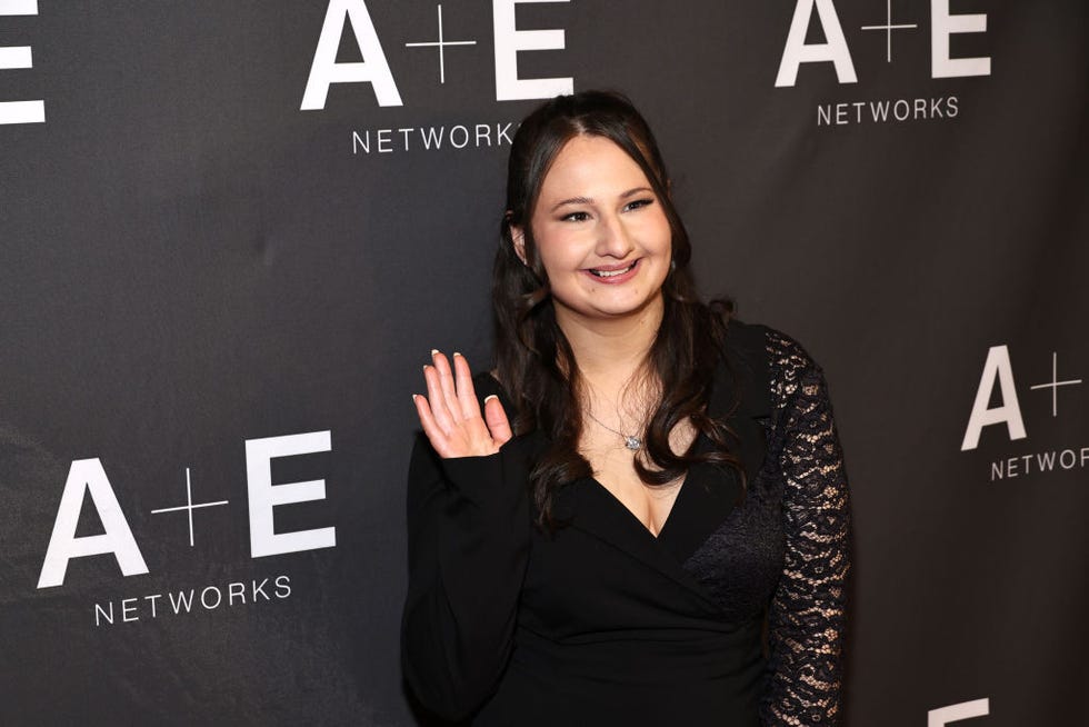 gypsy rose blanchard smiles and waves while standing in front of a black background with white logos, she wears a black long sleeve dress