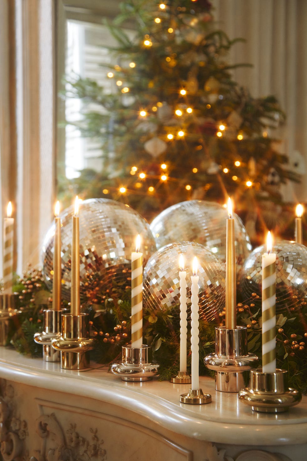 a festive arrangement features several lit candles in various holders, surrounded by greenery and glittering disco balls in the background, a decorated christmas tree twinkles with lights, creating a warm, inviting atmosphere the decorative elements include gold and white striped candles along with smooth metallic candle holders, adding a touch of elegance to the setting