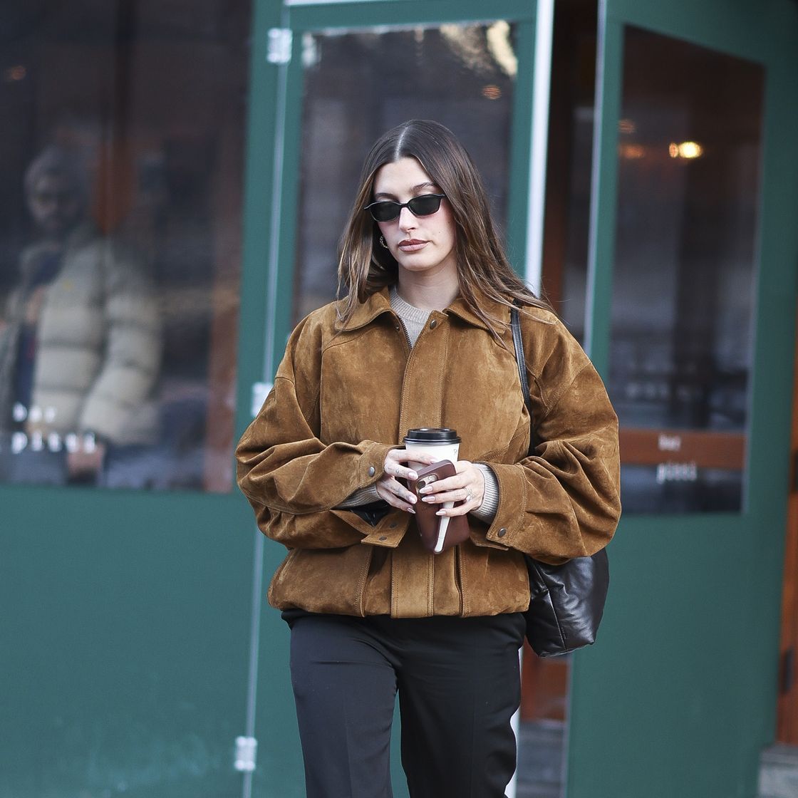 hailey bieber wearing a brown suede jacket whilst carrying a coffee and a rhode phone case