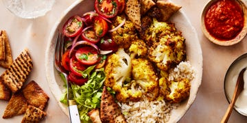 roasted cauliflower in a bowl with tomato salad, crushed pita, onions on top of rice and drizzled with white halal cart style sauce