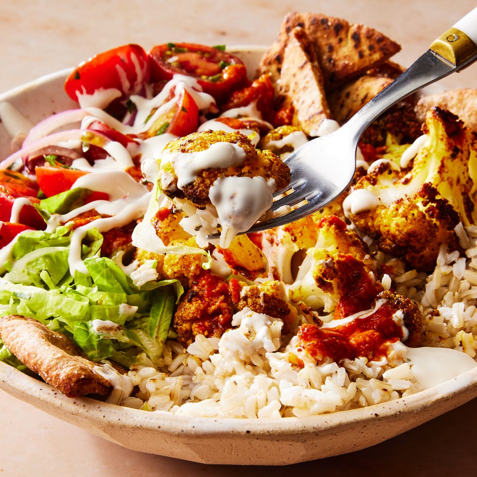 roasted cauliflower in a bowl with tomato salad, crushed pita, onions on top of rice and drizzled with white halal cart style sauce