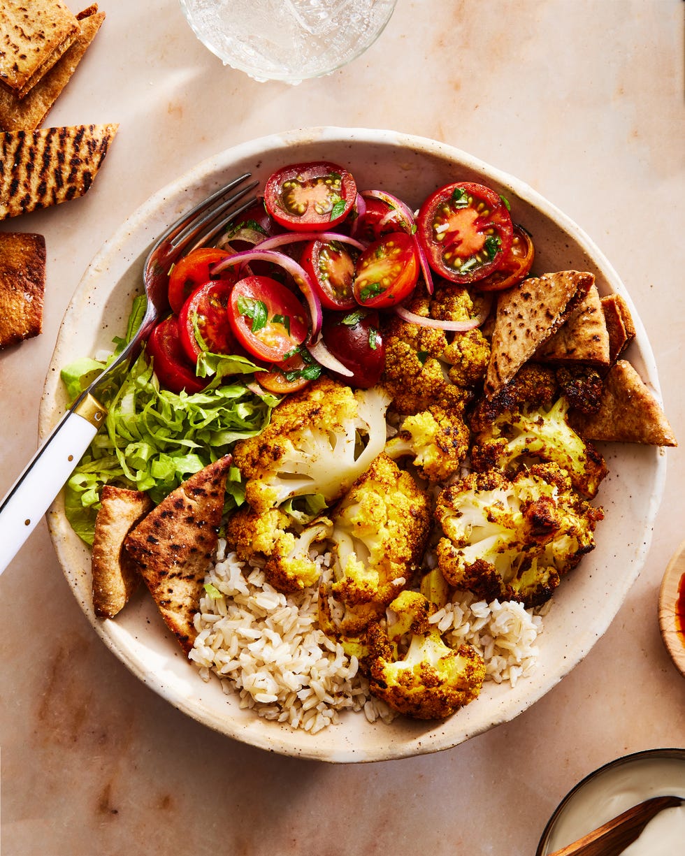 roasted cauliflower in a bowl with tomato salad, crushed pita, onions on top of rice and drizzled with white halal cart style sauce