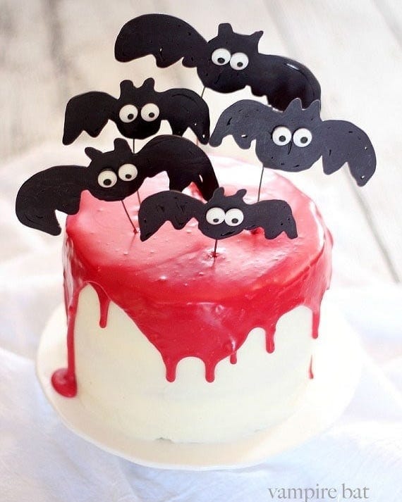 white frosted cake garnished with with red dripping ganache and vampire bat decorations