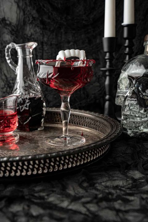 draculas kiss cocktail in a glass with a red candied rim and a pair of plastic vampire teeth perched on the rim