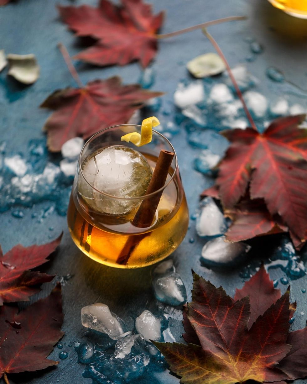smoked maple old fashioned in a glass with a cinnamon stick and a piece of curled lemon peel on a dark turquoise table covered in ice cubes and fall leaves