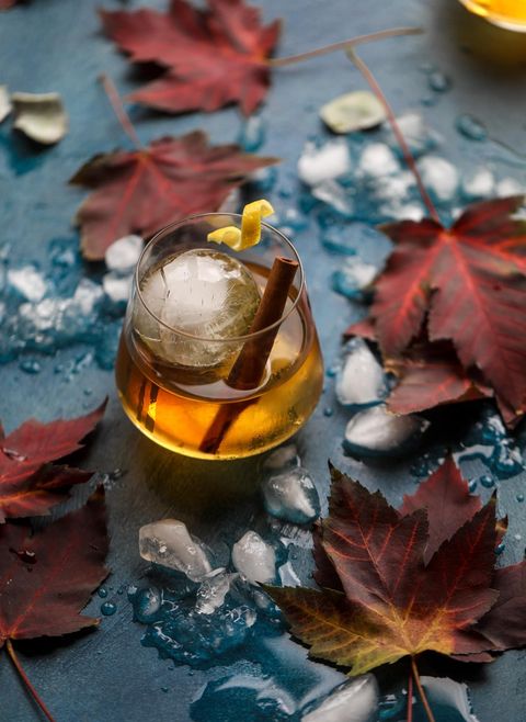 smoked maple old fashioned in a glass with a cinnamon stick and a piece of curled lemon peel on a dark turquoise table covered in ice cubes and fall leaves