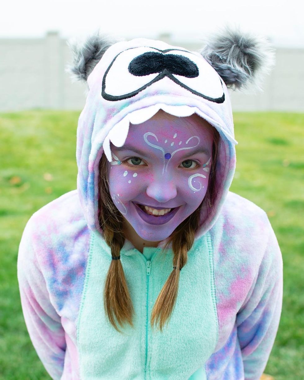 tween girl with braids wearing purple and green monster onesie pajamas with purple face paint for halloween costume