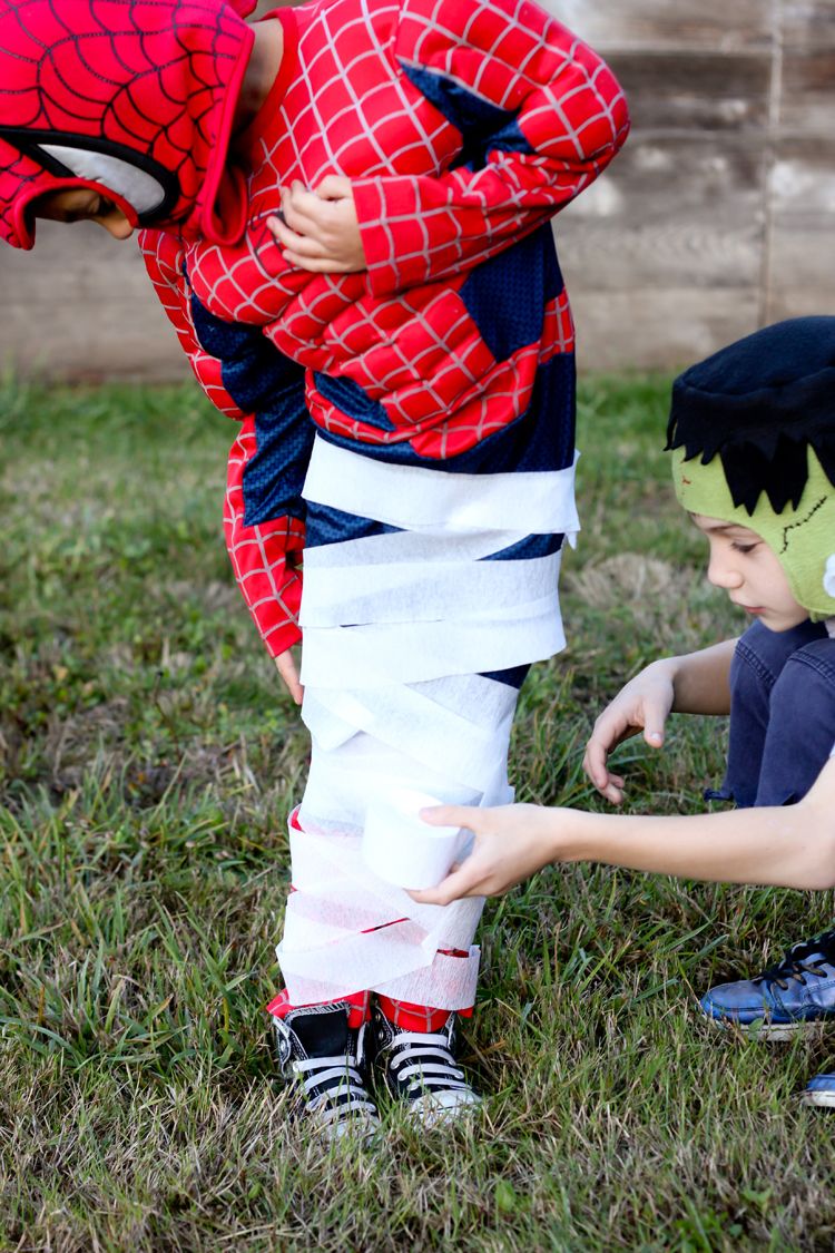 child wearing frankenstein costume wraps white crepe paper around legs of child wearing spiderman costume for mummy sack race