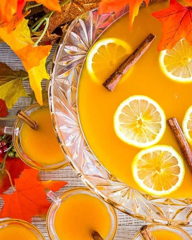 spiced pumpkin punch in punch bowl with lemons and cinnamon sticks glass mugs and leaves on side