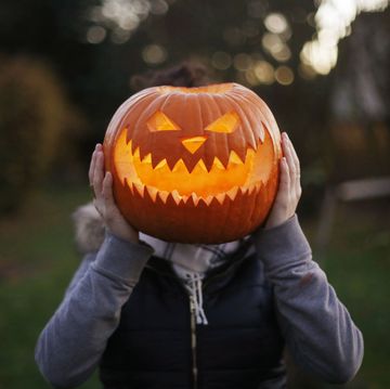 girl in zip up hoodie holding halloween jack o lantern in front of her head outdoors at dusk