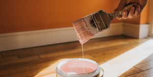 hand dipping a paint brush into a large tin of pink paint as it drips back into the pot