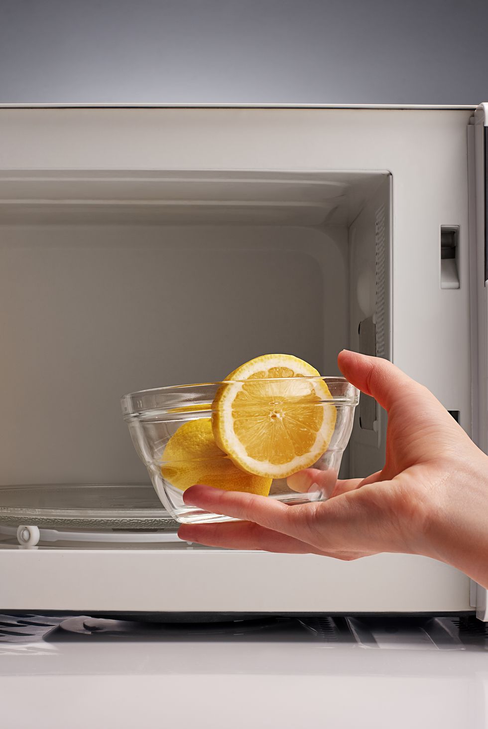 hand holding glass bowl with cut lemon in front of white open empty microwave on gray background