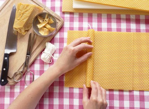 Hands carefully rolling beeswax sheet around candle, resting on red and white checked table cloth, wooden chopping board with knife, scissors, and string beside, above view.
