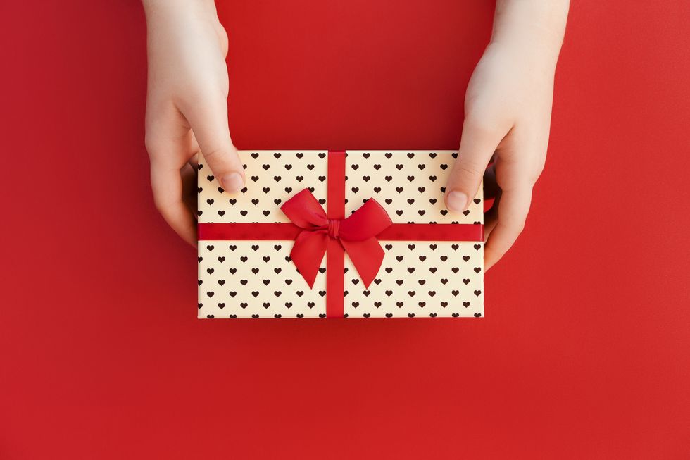 hands of teenage girl holding a beige gift box with hearts tied with a red satin ribbon on red background