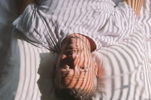 handsome adult man wakes up in the morning, lying on back, with sunlight illuminated him through window with blinds