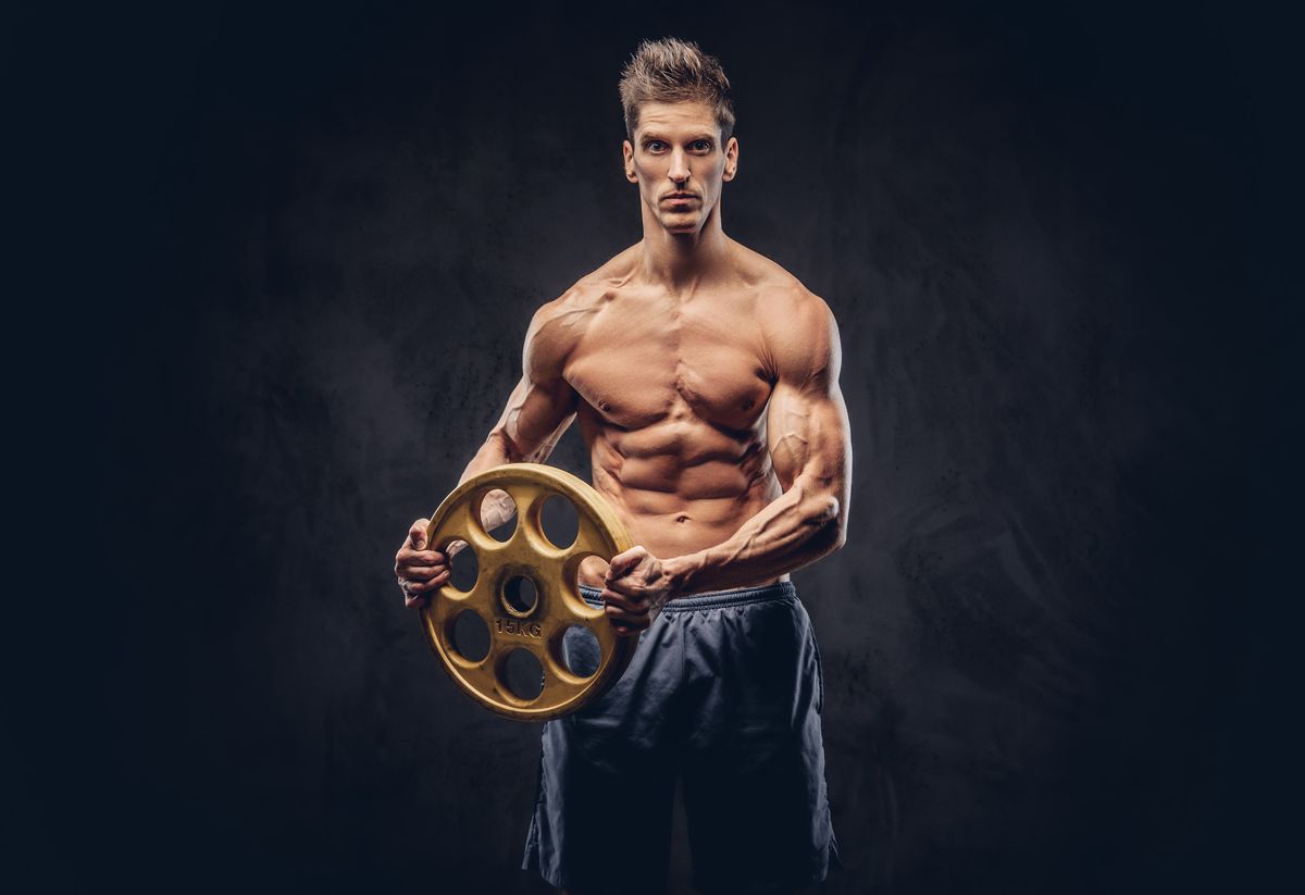 Handsome shirtless ectomorph bodybuilder with stylish hair posing with a barbell disk on dark background.