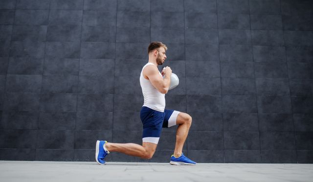 handsome strong muscular caucasian man in shorts and t shirt doing lunges and holding kettle bell in background is gray wall