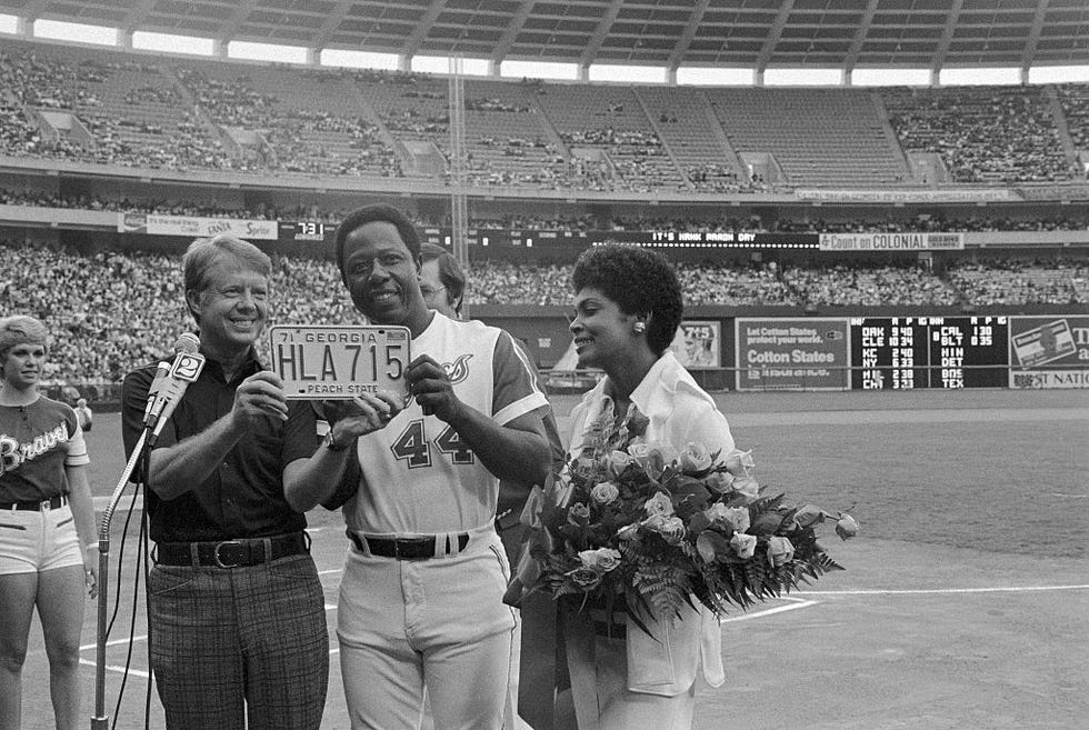georgia governor jimmy carter holds a specialized georgia license plate with baseball player hank aaron, the plate reads hla715, in front of carter is a microphone and to the right of aaron is his wife holding a large bouquet of roses and greenery, they are standing on the baseball field at a stadium that is visible in the background