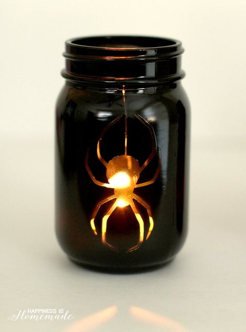 a mason jar painted black, the black is cut out in the shape of a dangling spider, through which a candle is visible