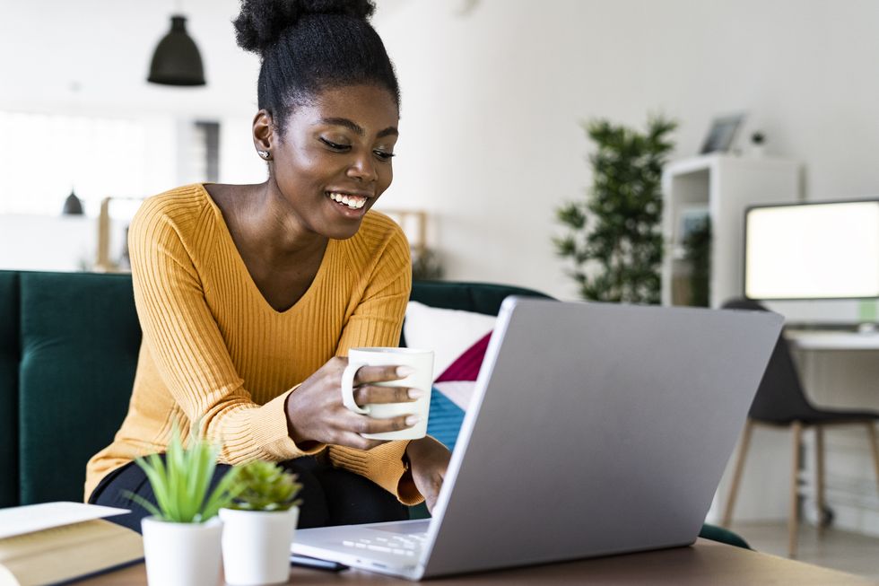 happy afro woman holding cup while using laptop at home