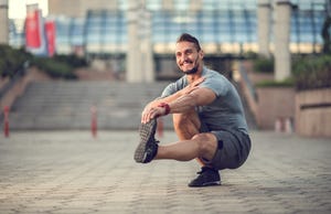 Happy athletic man stretching his leg while crouching outdoors.