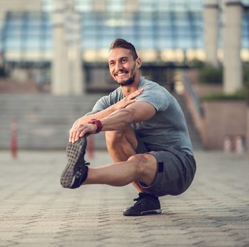 Happy athletic man stretching his leg while crouching outdoors.