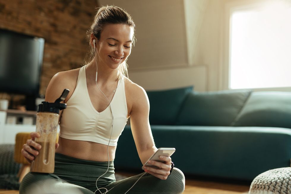 happy athletic woman using mobile phone while drinking smoothie at home