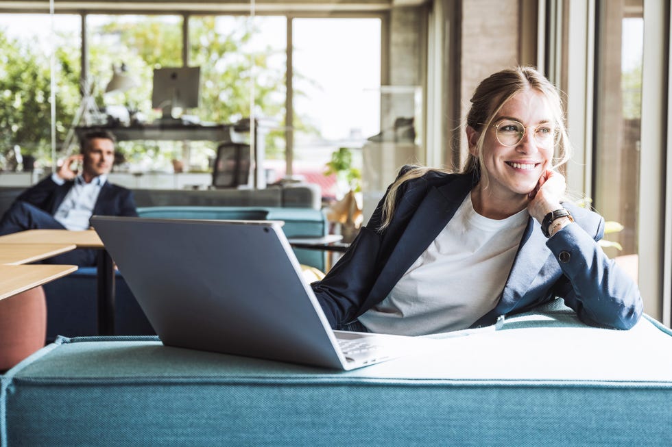 happy businesswoman sitting with laptop on sofa in office