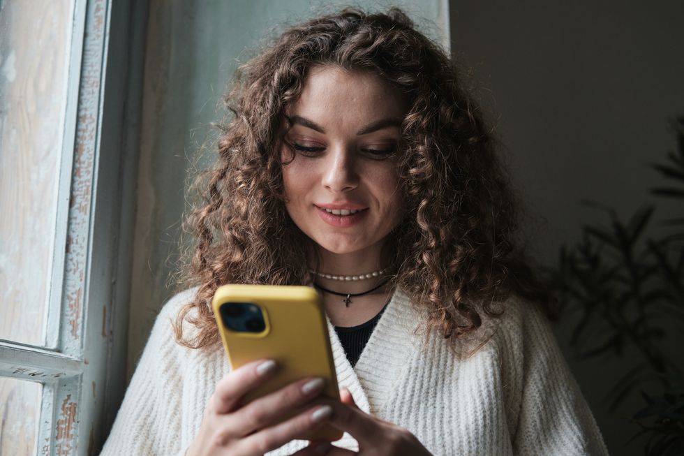 happy curly woman using smartphone at home, smiling girl holding mobile phone chatting with friends online