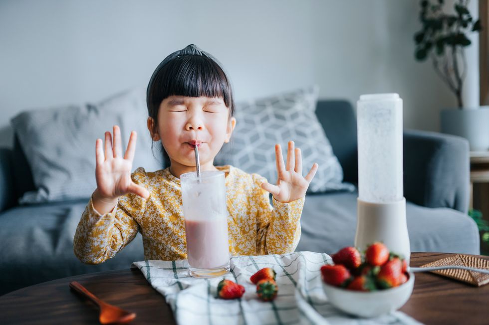 happy little asian girl drinking a glass of healthy homemade strawberry smoothie at home she is excited with her hands raised and eyes closed while drinking her favourite smoothie healthy eating and healthy lifestyle concept