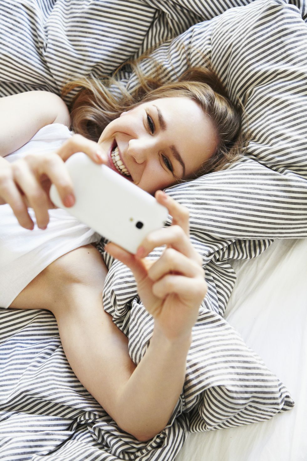 quarantine birthday ideas - happy mid-adult woman laying in bed taking a selfie with her smartphone