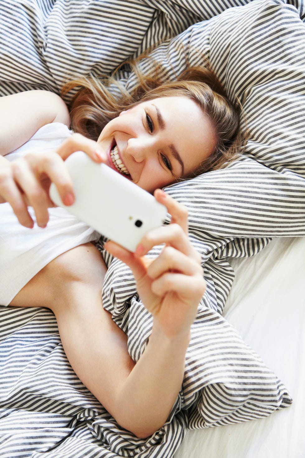 quarantine birthday ideas - happy mid-adult woman laying in bed taking a selfie with her smartphone