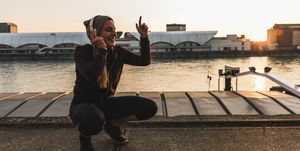 Happy sportive young woman listening to music at the riverside in the city at sunset