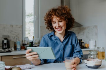 happy woman drinking coffee, eating breakfast and reading something on a digital tablet in her kitchen in the morning