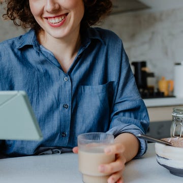 happy woman drinking coffee, eating breakfast and reading something on a digital tablet in her kitchen in the morning