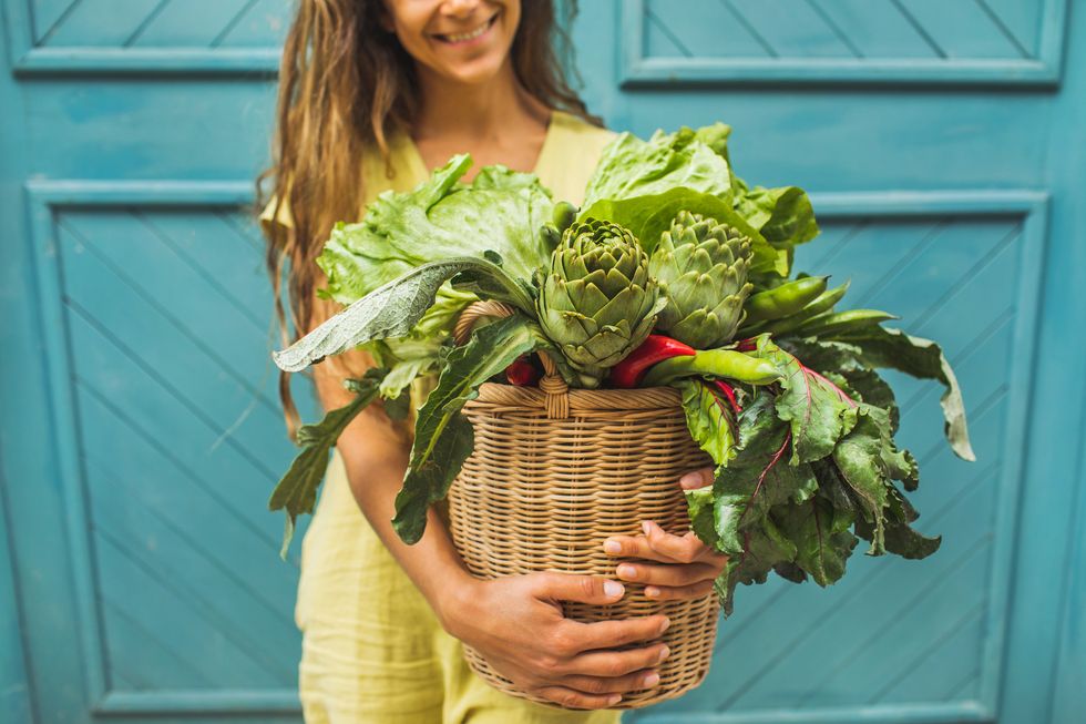 happy woman holding wicker basket with fresh greens and vegetables against blue door background with unrecognizable person