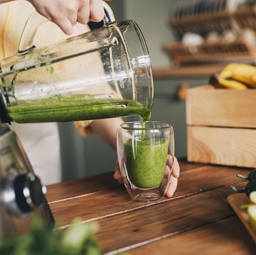 happy woman on detox diet, pouring green cocktail from mixer into glass in kitchen