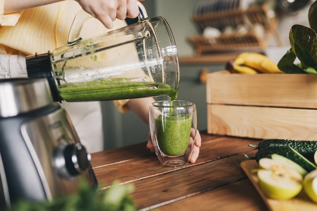 happy woman on detox diet, pouring green cocktail from mixer into glass in kitchen
