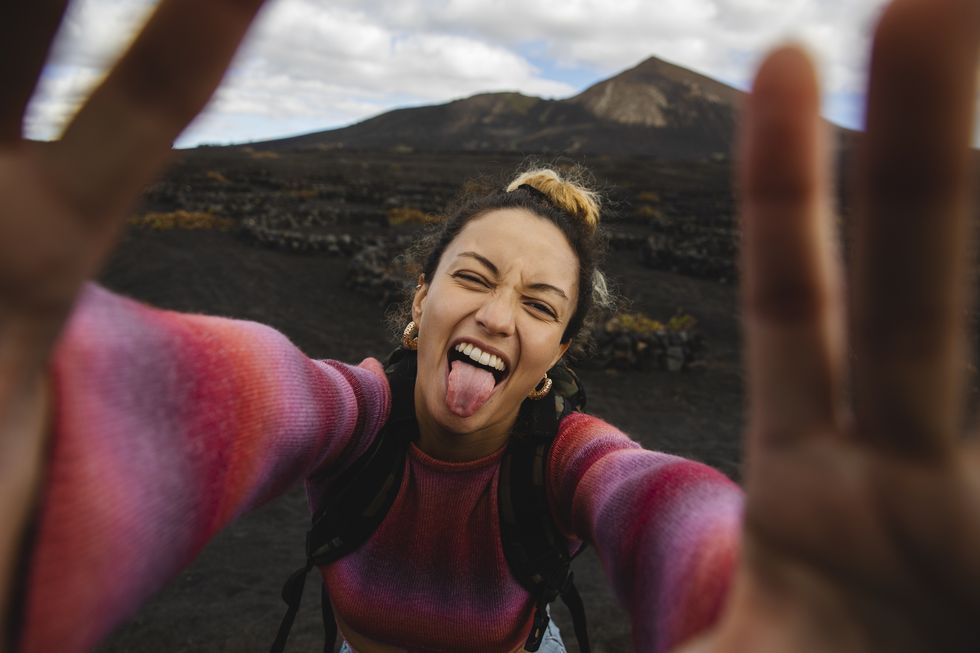 happy woman sticking out tongue and taking selfie with landscape in background
