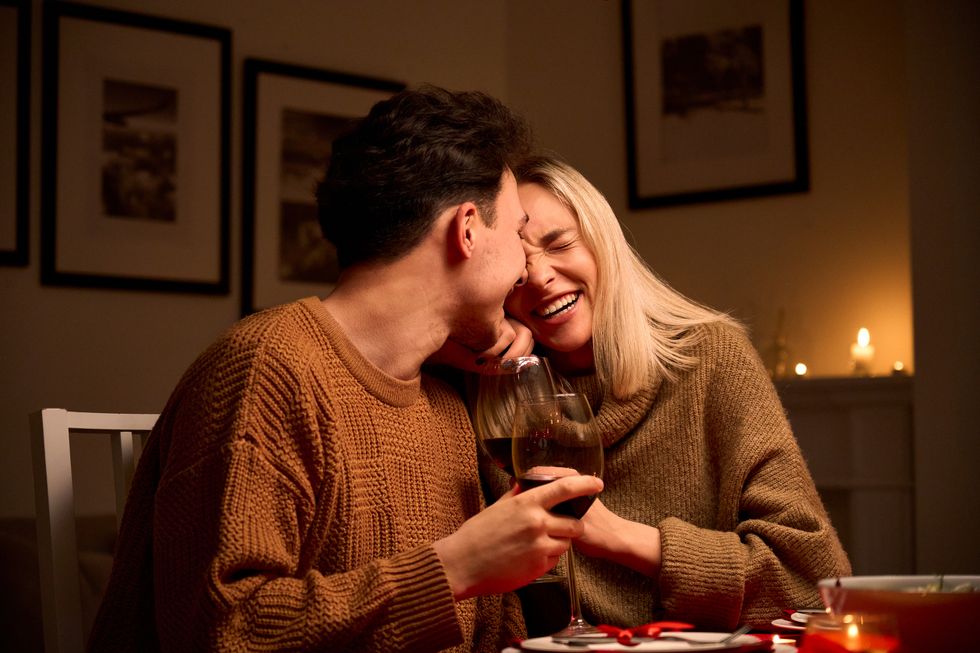 happy young couple in love hugging, laughing, drinking wine, enjoying talking, having fun together celebrating valentines day dining at home, having romantic dinner date with candles sitting at table