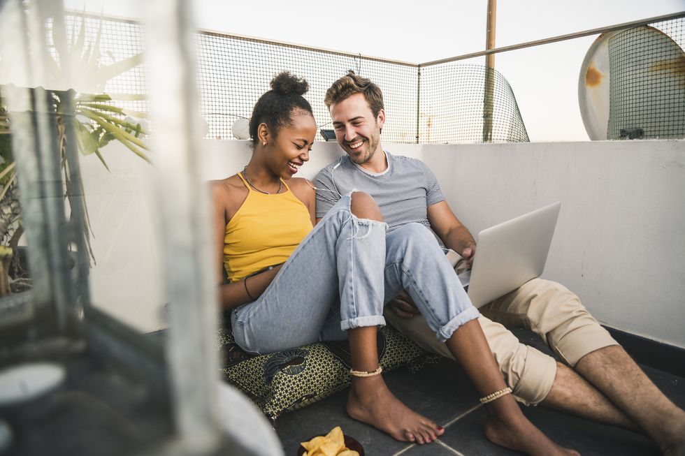 happy young couple with laptop and earphones sitting on rooftop in the evening