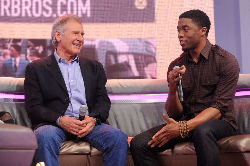 harrison ford and chad boseman sit on a couch, boseman talks into a microphone he holds, ford looks at boseman smiling and holds a microphone in his lap