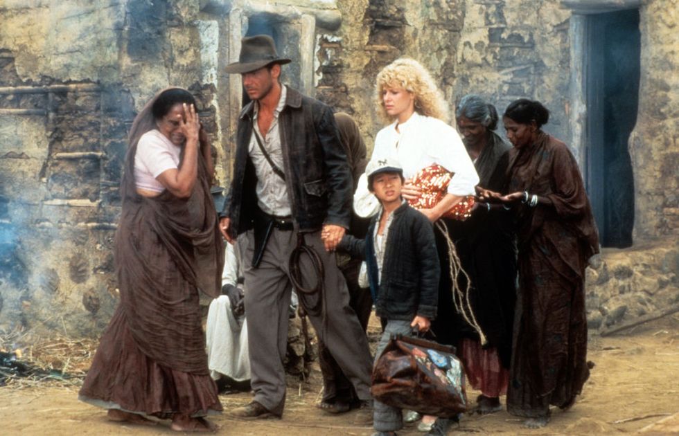 harrison ford, ke huy quan, and kate capshaw on the movie set for indiana jones and the temple of doom, walking past a stone building and several women dressed in brown clothes