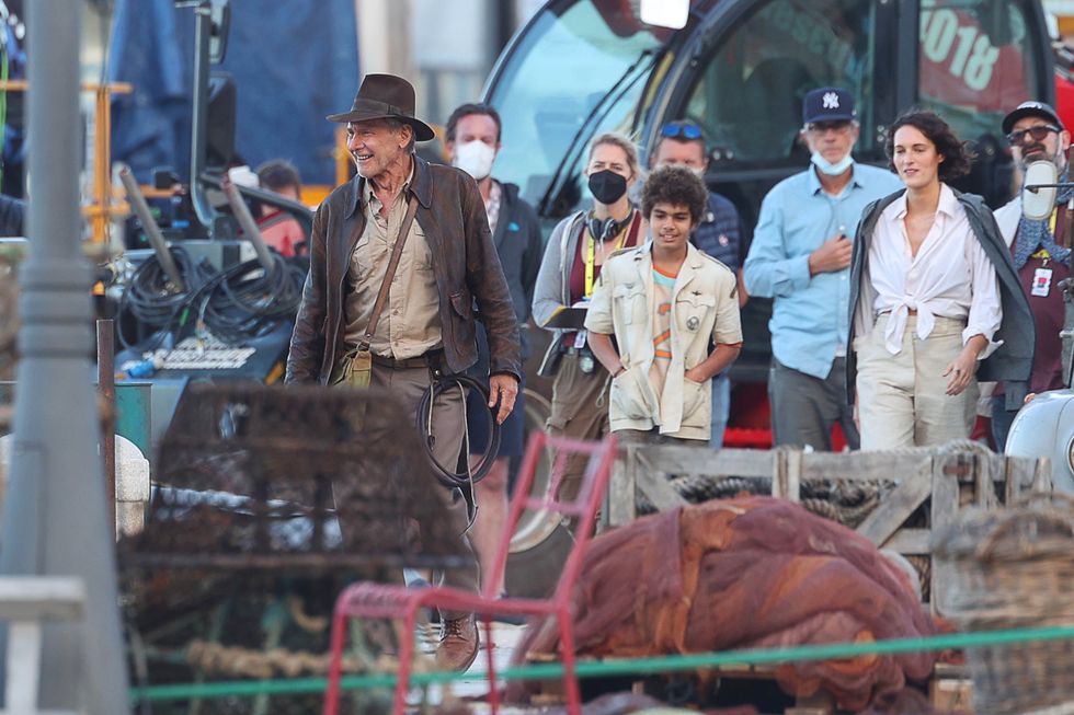 harrison ford, dressed like indiana jones, smiling on a film set among a group of other people, including actress phoebe waller bridge wearing a white shirt and cream pants