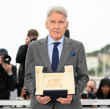 harrison ford, wearing a gray suit and blue tie, holds the palm d or award on a stage while photographer behind him take pictures